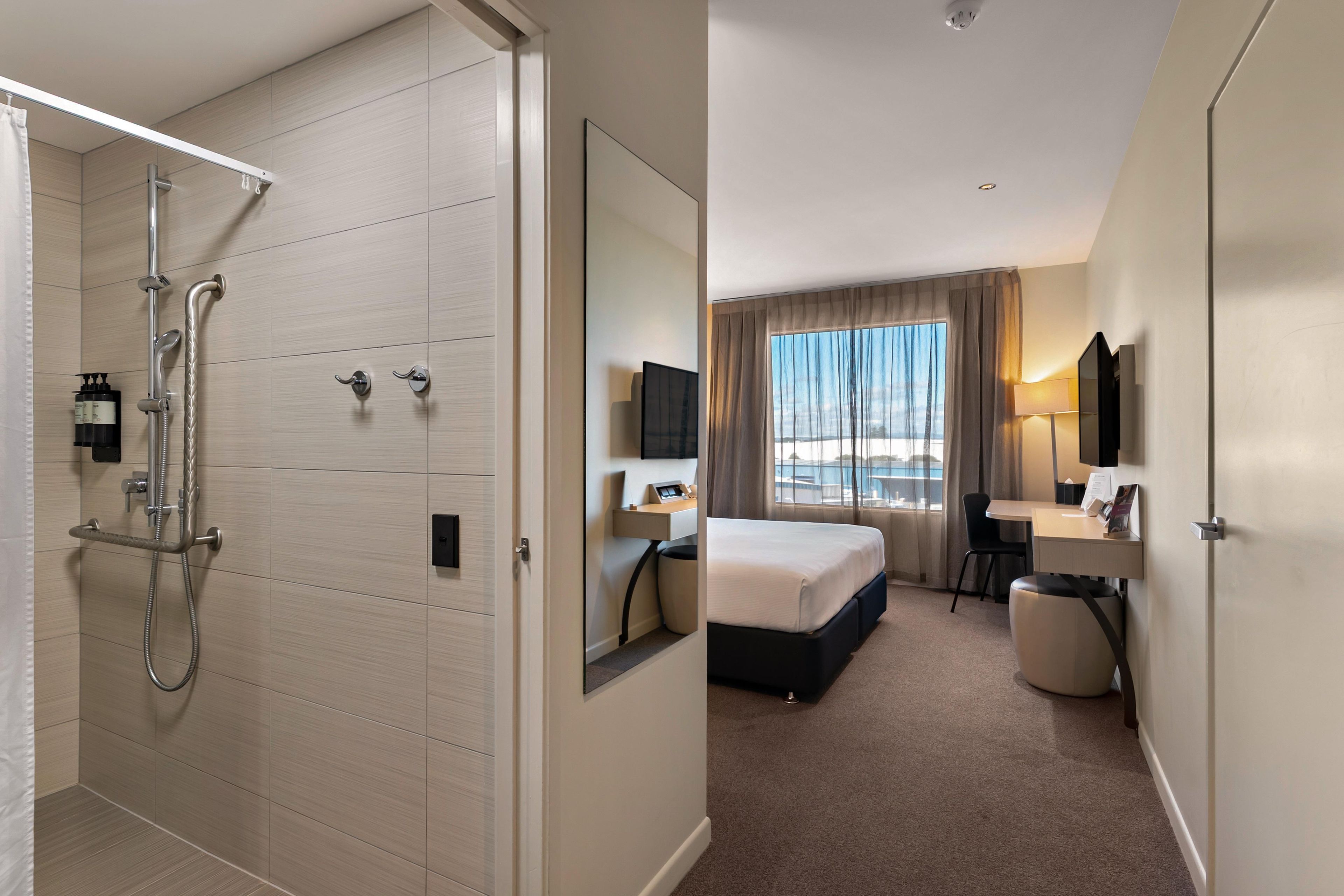 A hotel room with a walk-in shower, a bed, a desk, a chair, a mirror, and a window with a view.