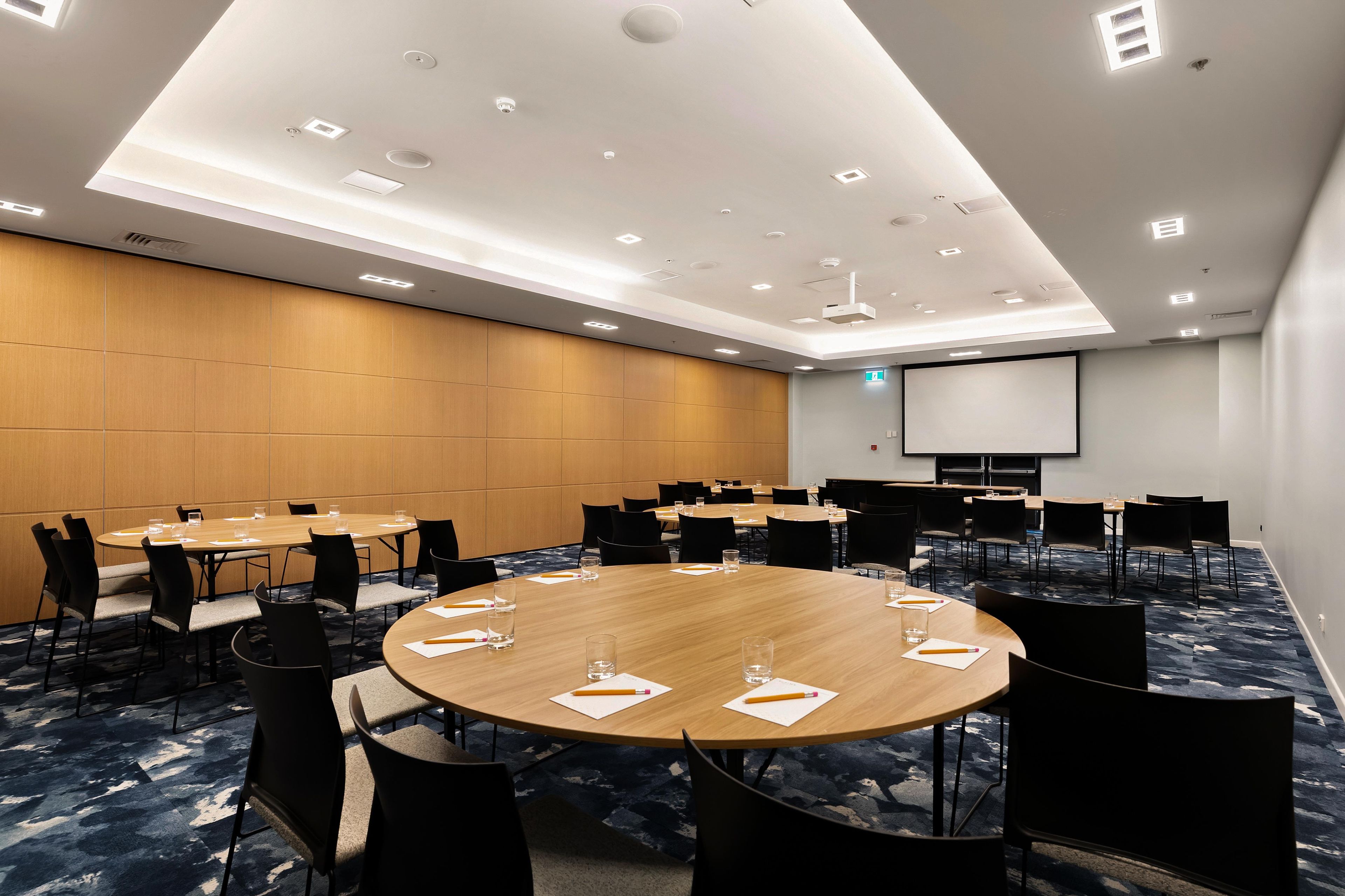 Un salón de conferencias con varias mesas y sillas, una pantalla de proyector e iluminación empotrada en el techo.