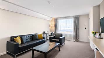 Quarto de hotel moderno com sofá de couro preto, mesa de centro, escrivaninha e uma janela grande com cortinas.