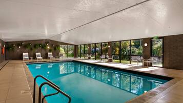 Une piscine intérieure aux parois en verre transparent, entourée de chaises longues et de tables, et offrant une vue sur la verdure environnante.