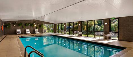 An indoor swimming pool with clear glass walls, surrounded by lounge chairs and tables, and a view of greenery outside.