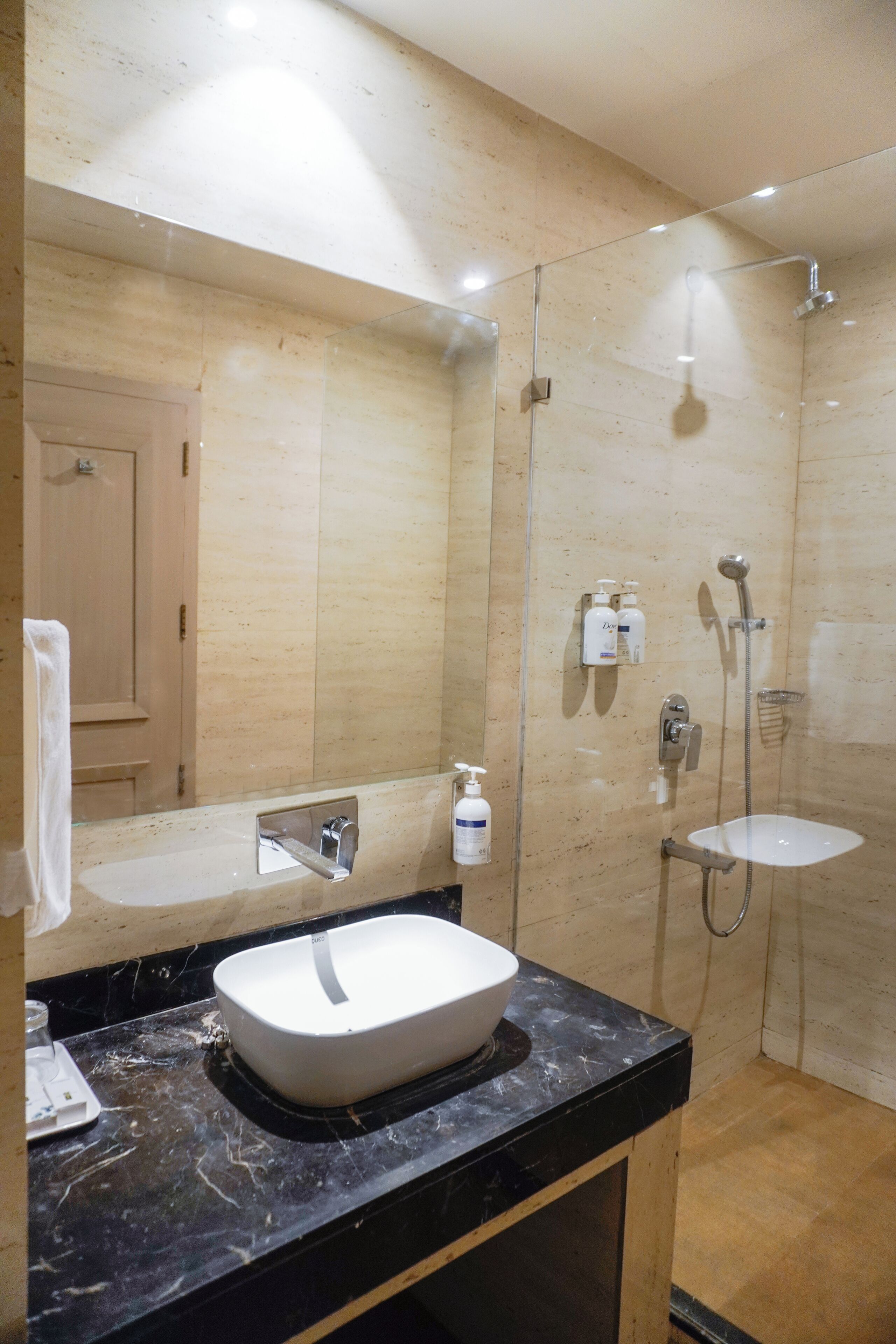 Une salle de bain moderne avec un grand miroir, un comptoir en marbre noir, un lavabo blanc, une douche à parois de verre et une porte en bois à l’arrière-plan.