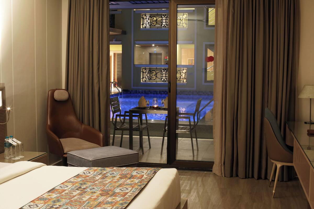 A hotel room with a bed, a desk, a chair, and a view of an indoor swimming pool through a glass door.