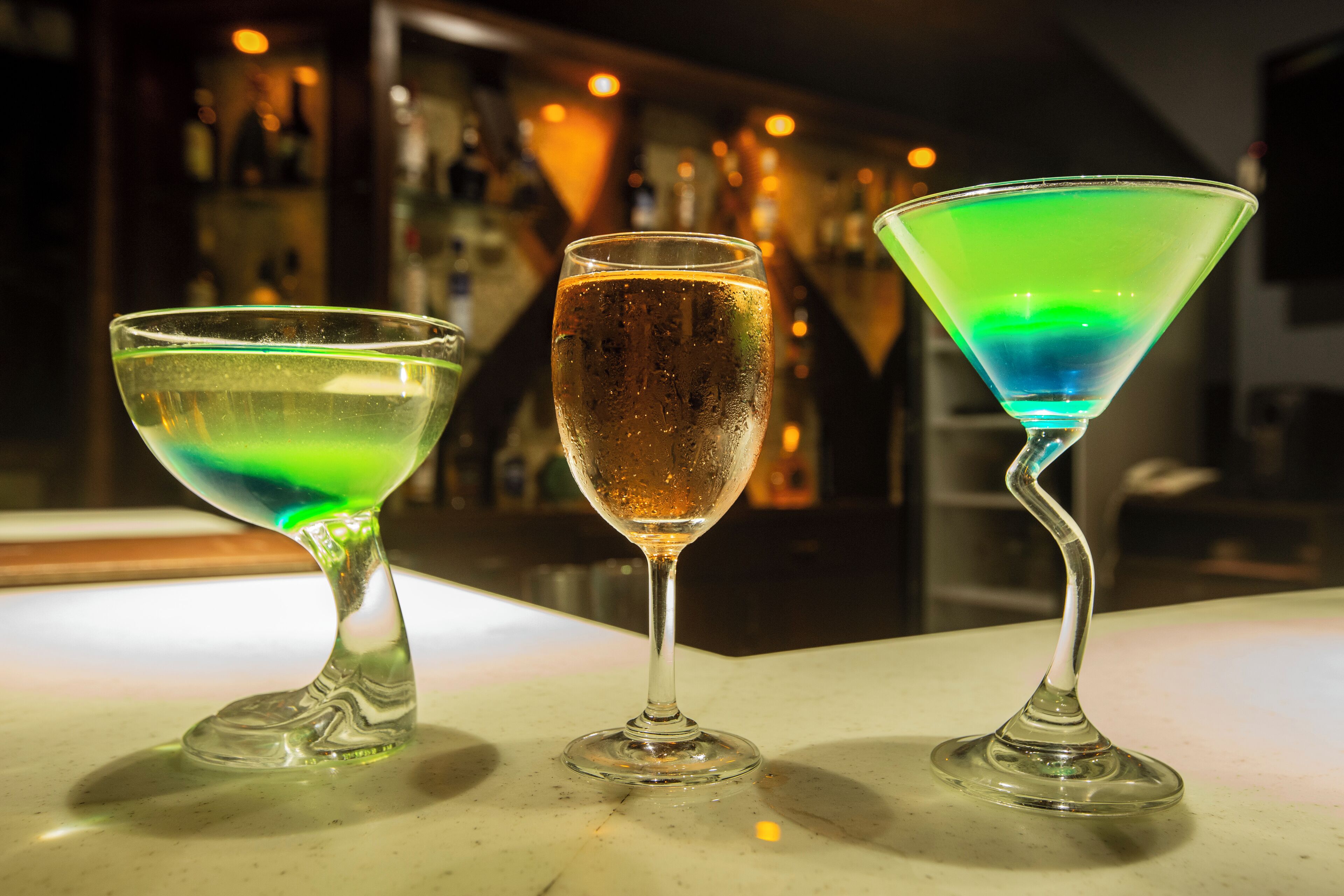 Tres vasos de cócteles de colores distintos sobre una barra.