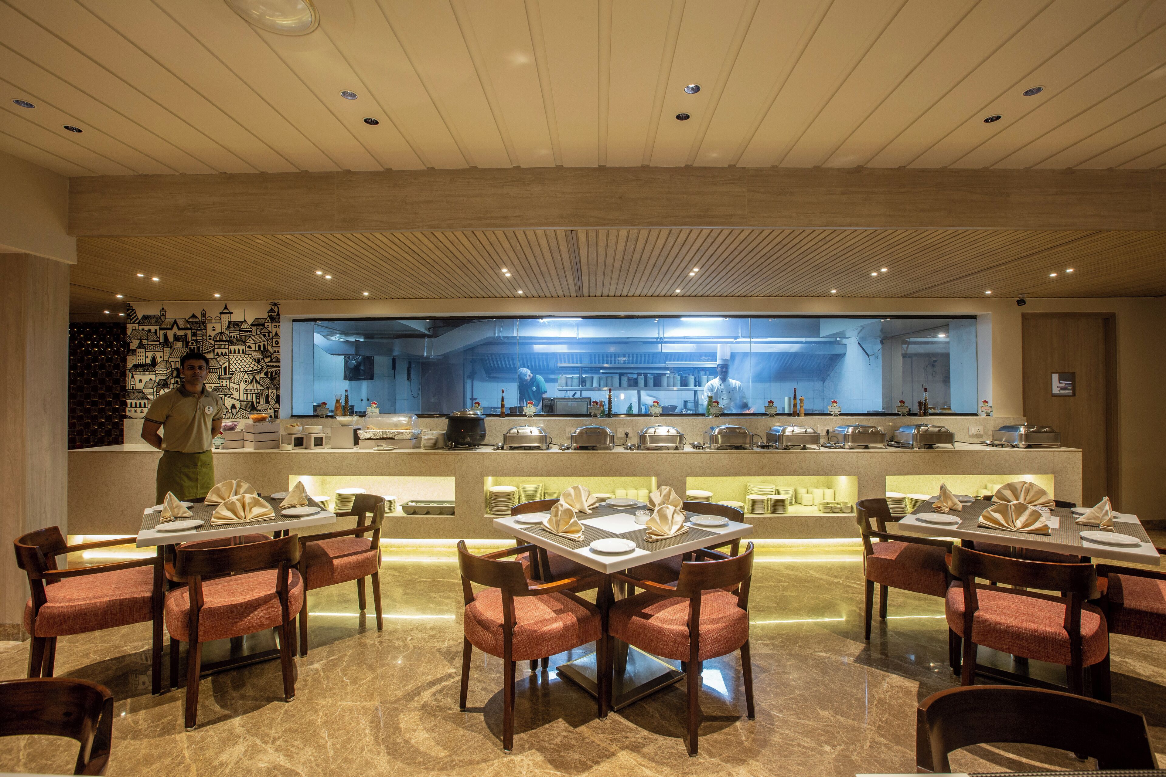 A restaurant with a modern interior, featuring a central counter with a view of the kitchen, wooden tables with red cushioned chairs, and a polished marble floor.