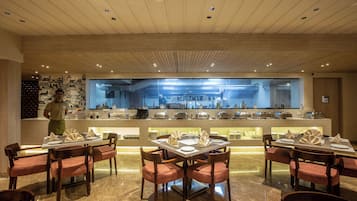 A restaurant with a modern interior, featuring a central counter with a view of the kitchen, wooden tables with red cushioned chairs, and a polished marble floor.