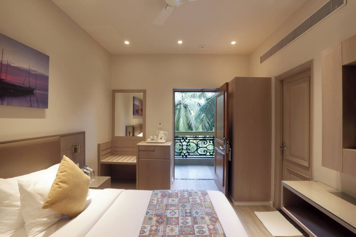A hotel room with a bed, a nightstand, a desk, a mirror, and a balcony with a view of palm trees.