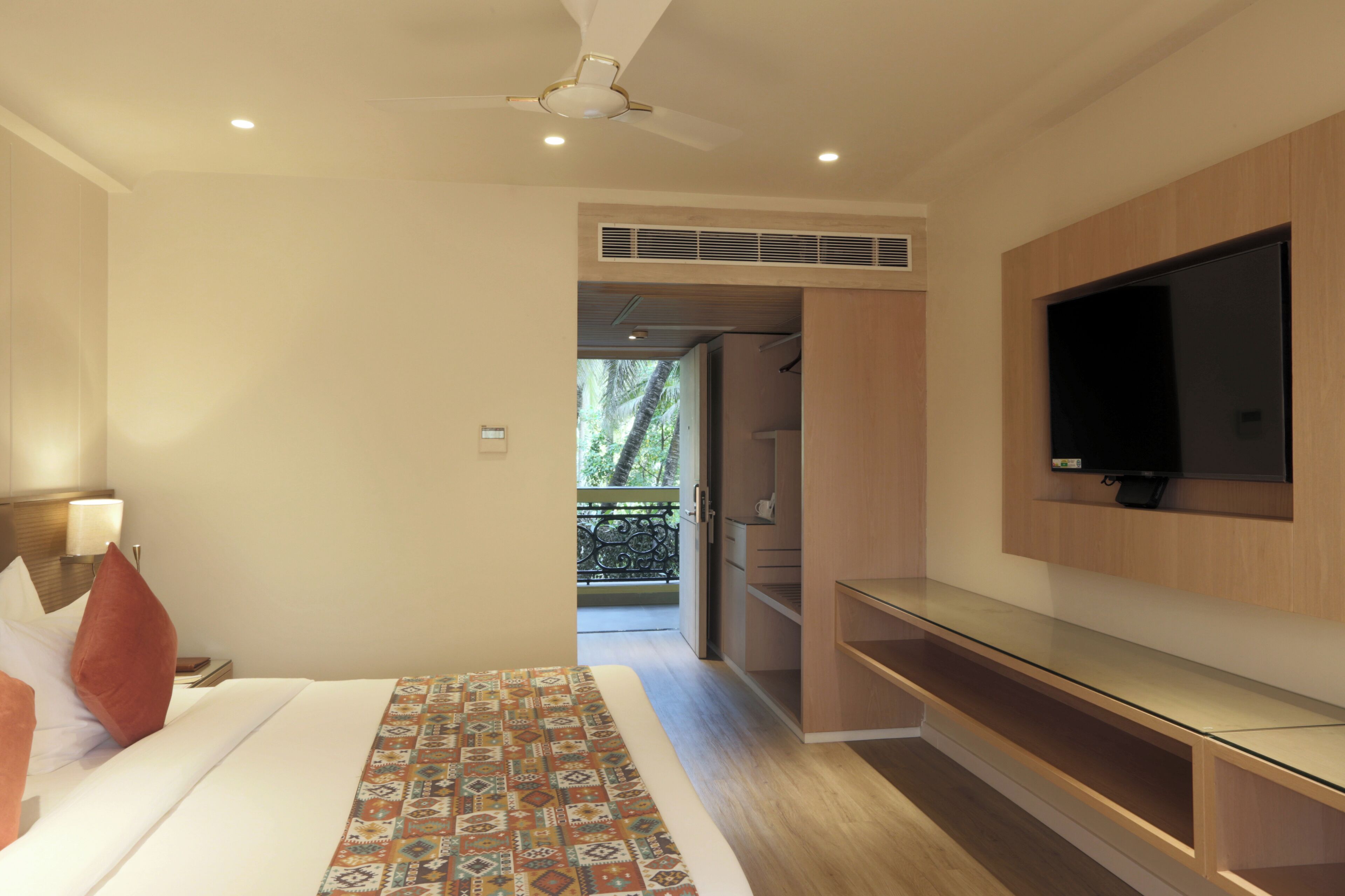 A modern hotel room with a bed, a TV mounted on the wall, and a balcony with a view of palm trees.