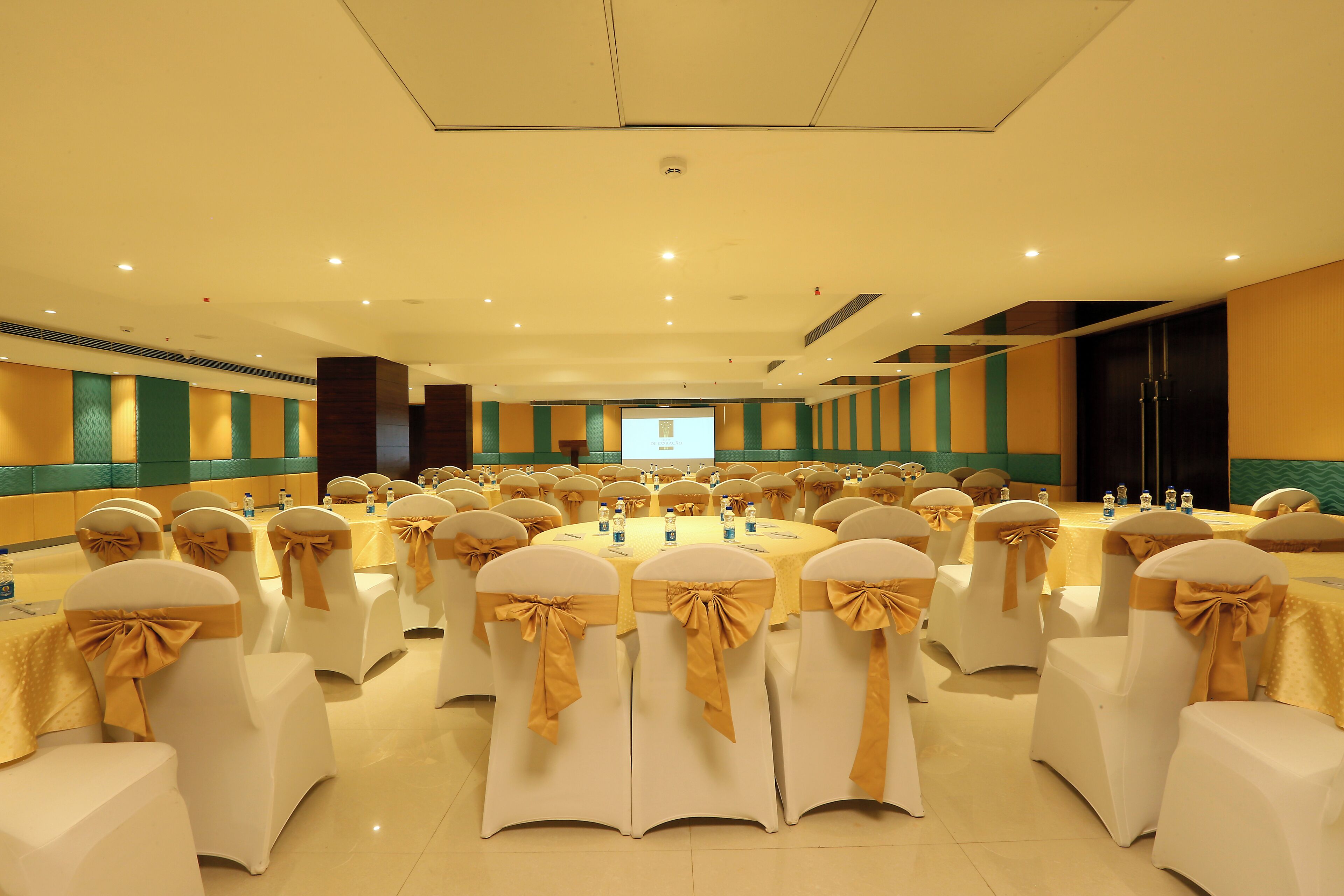 A conference room with round tables, white chairs with gold sashes, and a projection screen at the front.