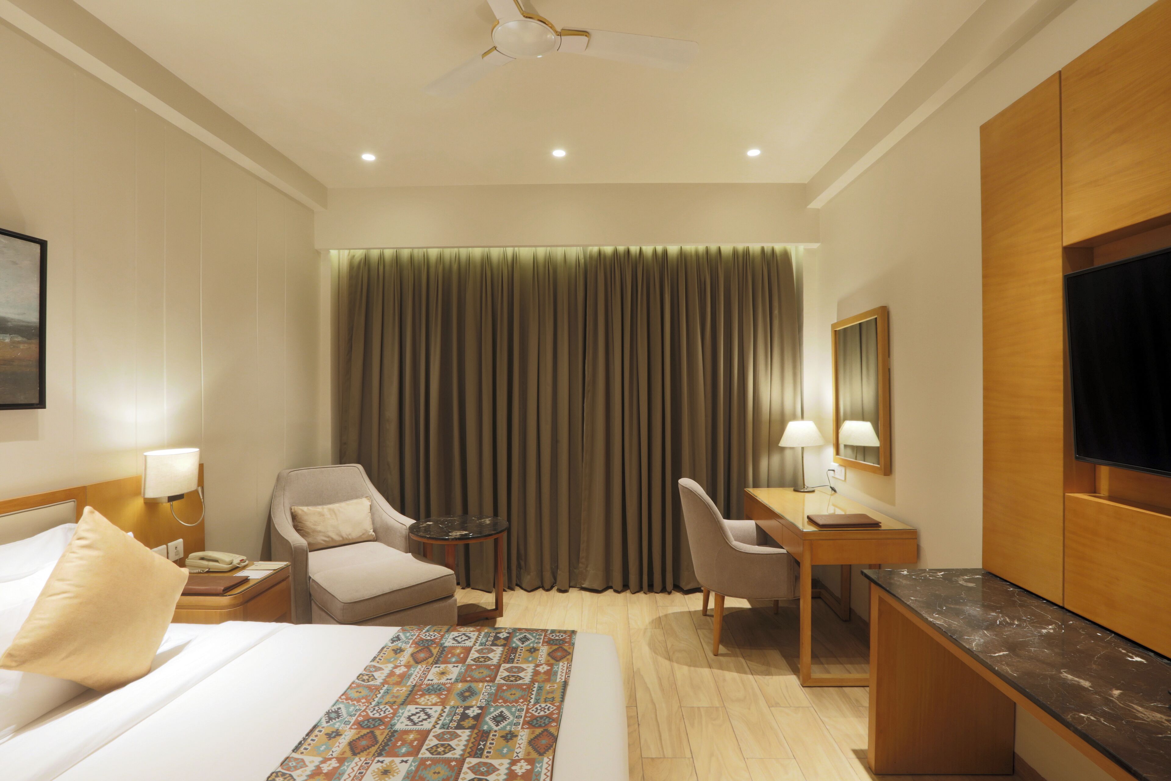 Une chambre d’hôtel comprenant un lit, deux fauteuils, un bureau avec une chaise, une télévision et un ventilateur de plafond.