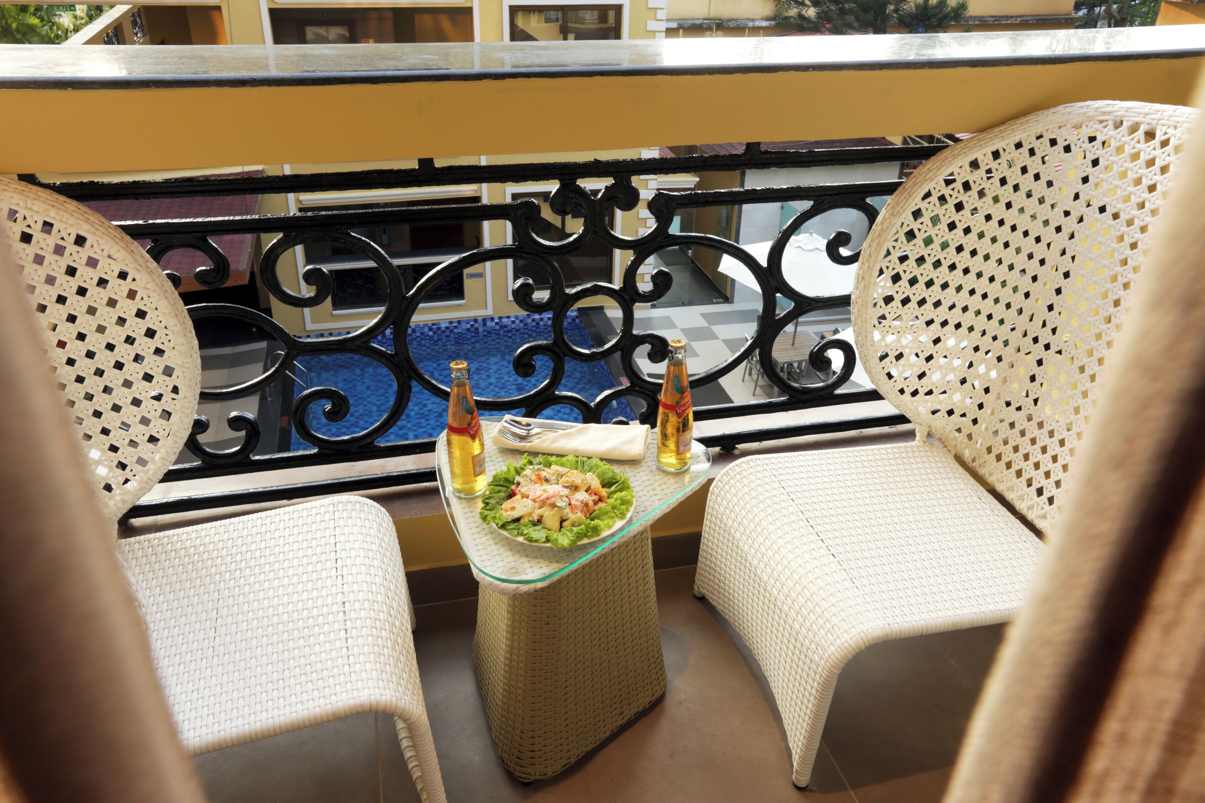 A balcony with two wicker chairs, a glass table with a salad, and two bottles of wine.