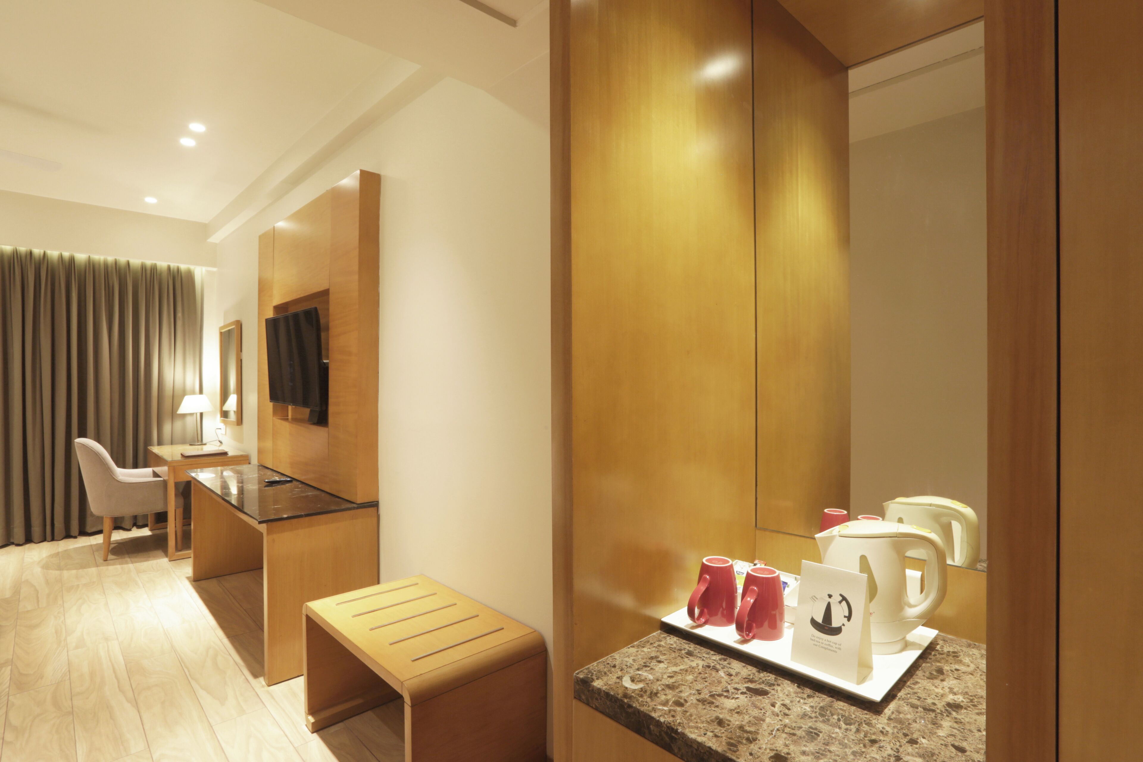 A modern hotel room with a wooden desk, a small stool, a mirror, and a kitchenette area with a coffee maker and cups.