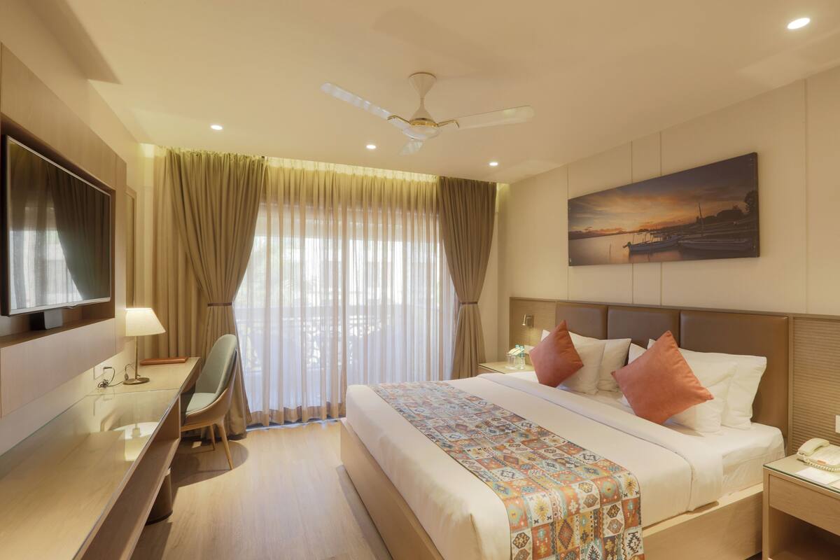 A hotel room with a bed, a desk, a chair, a TV, a painting on the wall, and a ceiling fan.