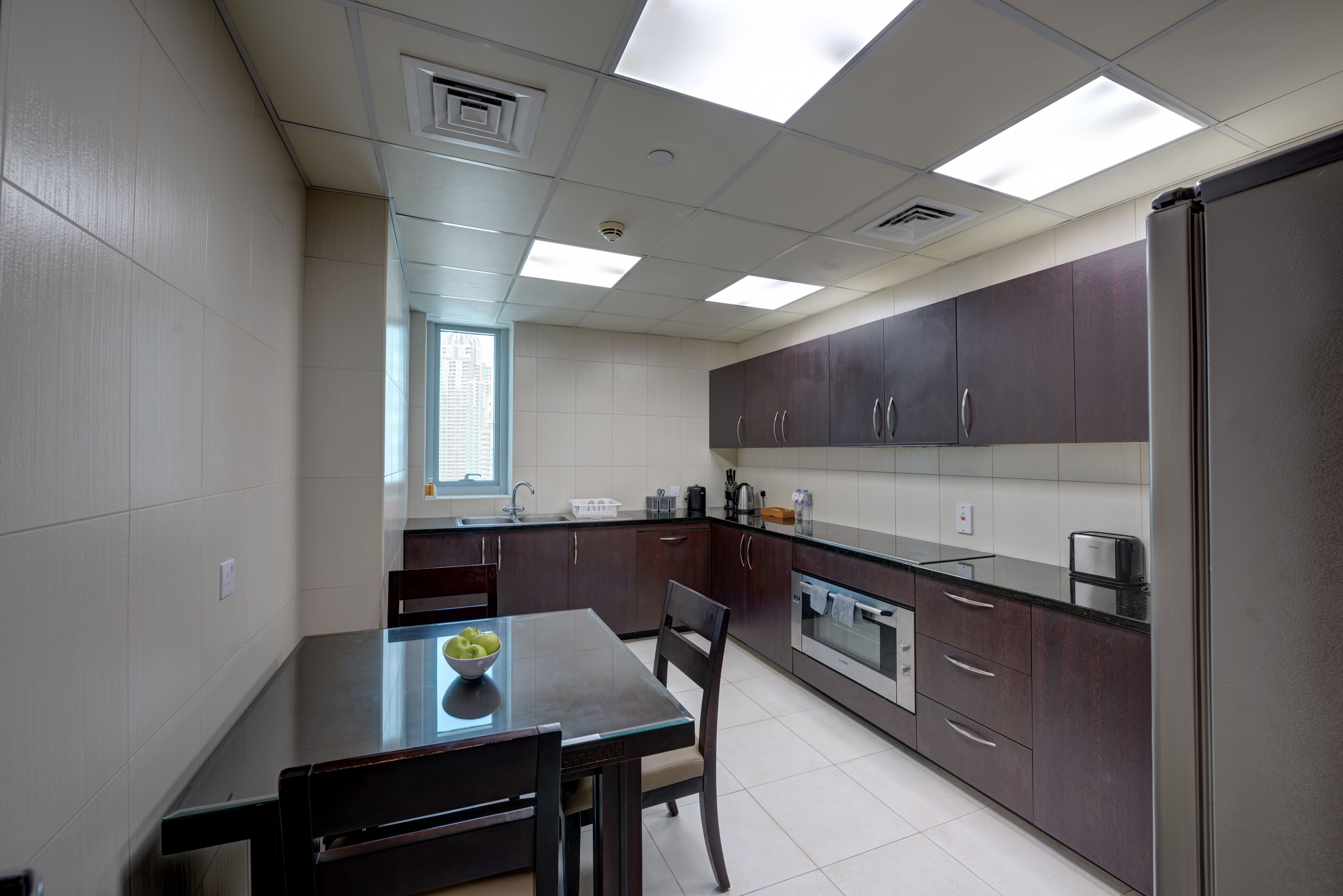A modern kitchen with dark cabinets, a stainless steel refrigerator, and a glass dining table.