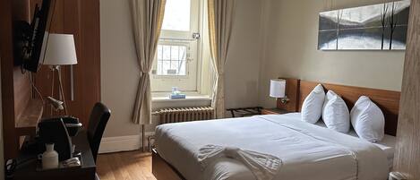 A hotel room with a large bed, a desk with a lamp, a chair, a window with curtains, and a painting on the wall.