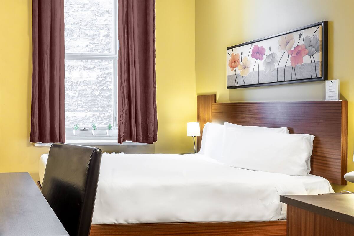 A hotel room with a bed, a desk, a chair, a window with curtains, a painting on the wall, and a bedside lamp.