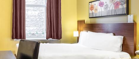 A hotel room with a bed, a desk, a chair, a window with curtains, a painting on the wall, and a bedside lamp.