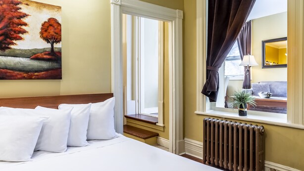 A hotel room with a bed, white pillows, a painting of trees, a window with curtains, a radiator, and a view of a plant and a couch. - Hotel Ermitage (Québec City)