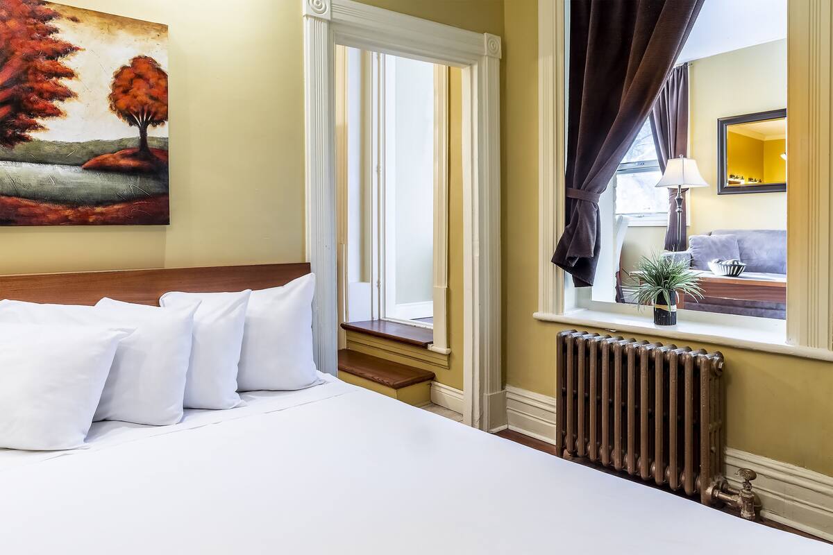 A hotel room with a bed, pillows, a window with curtains, a radiator, and a painting on the wall.