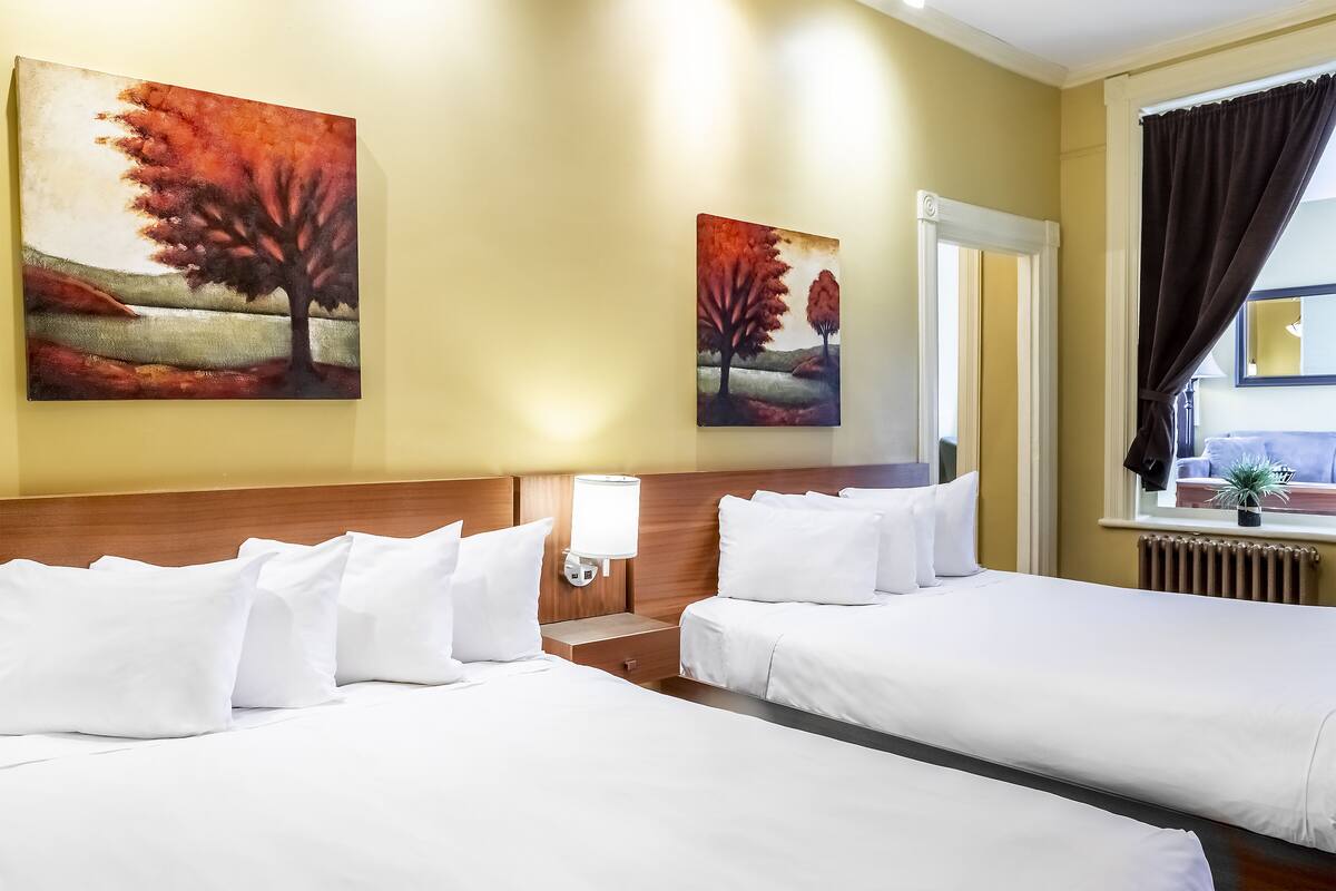 A hotel room with two beds, white bedding, a nightstand with a lamp, and two paintings on the wall.