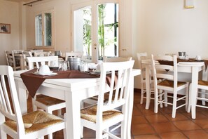 A dining area with white tables and chairs, some with woven seat cushions. There are metal buckets and a white tablecloth on one table.