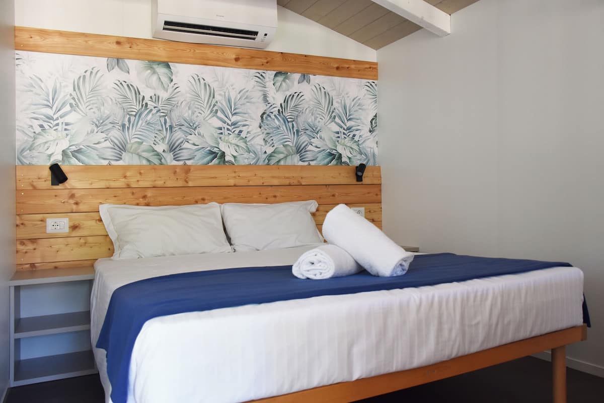 Un lit bien fait, recouvert d’une courtepointe bleue et blanche, avec des oreillers blancs et une serviette blanche enroulée. La tête de lit est en bois et recouverte d’un papier peint à motifs floraux.