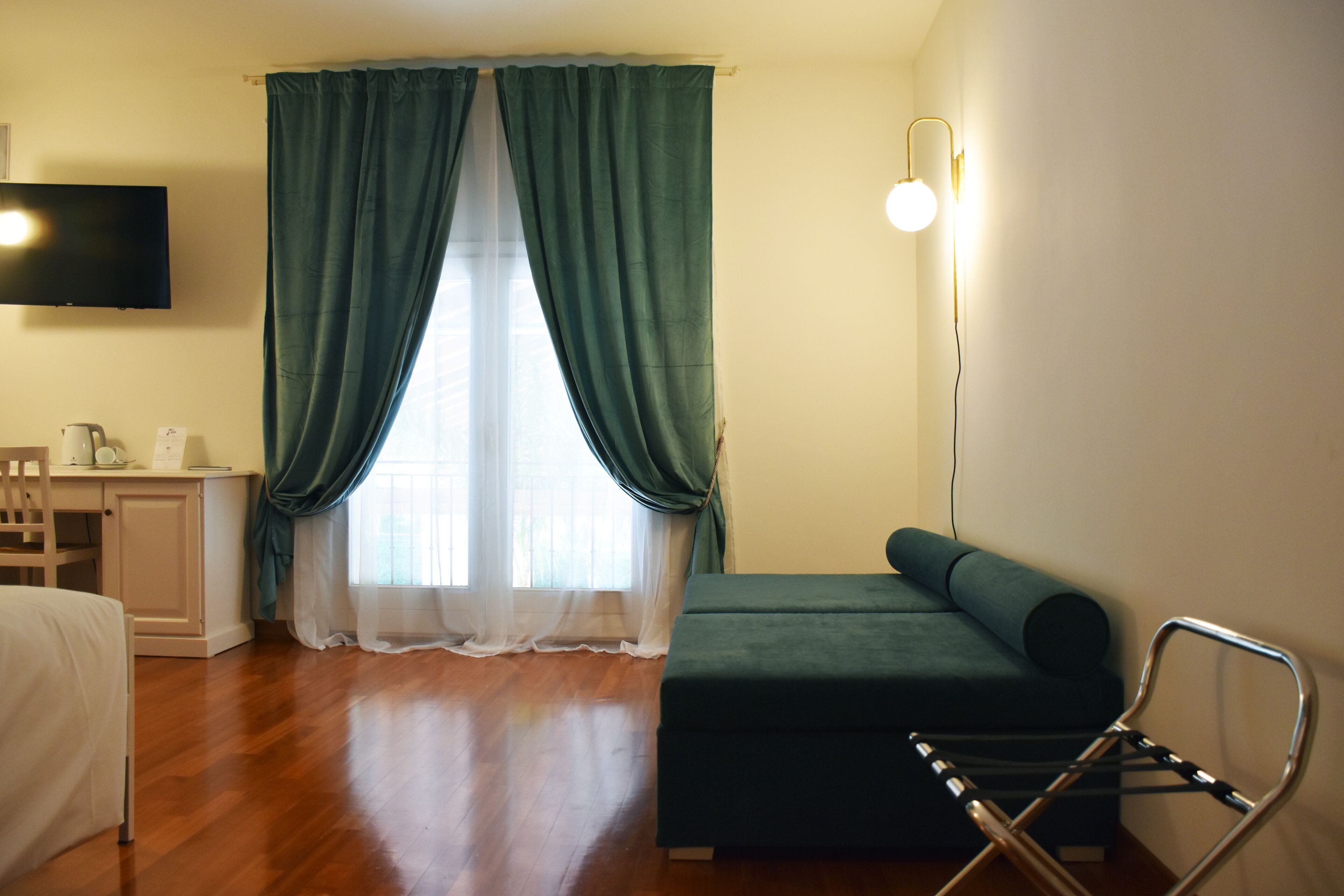 Una habitación con una cama, un escritorio con una silla, un sofá verde, una lámpara de pie y un televisor en la pared.