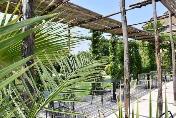 A covered outdoor seating area with tables and chairs, surrounded by greenery and trees.