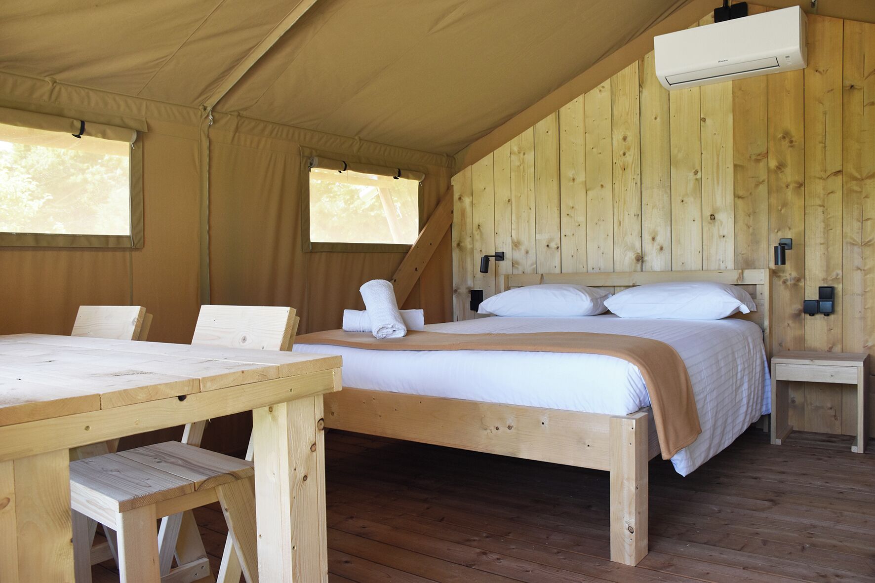 A tented accommodation with a wooden interior, a bed with white bedding, a table with chairs, and a wall-mounted air conditioning unit.