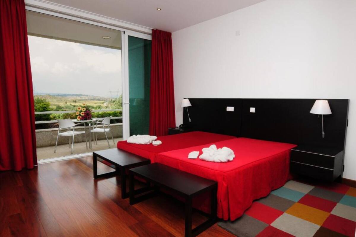 Ein Hotelzimmer mit einem großen Bett, zwei Nachttischen, einem Schreibtisch und einem Sessel. Es gibt einen Balkon mit roten Vorhängen und Blick auf die Landschaft.