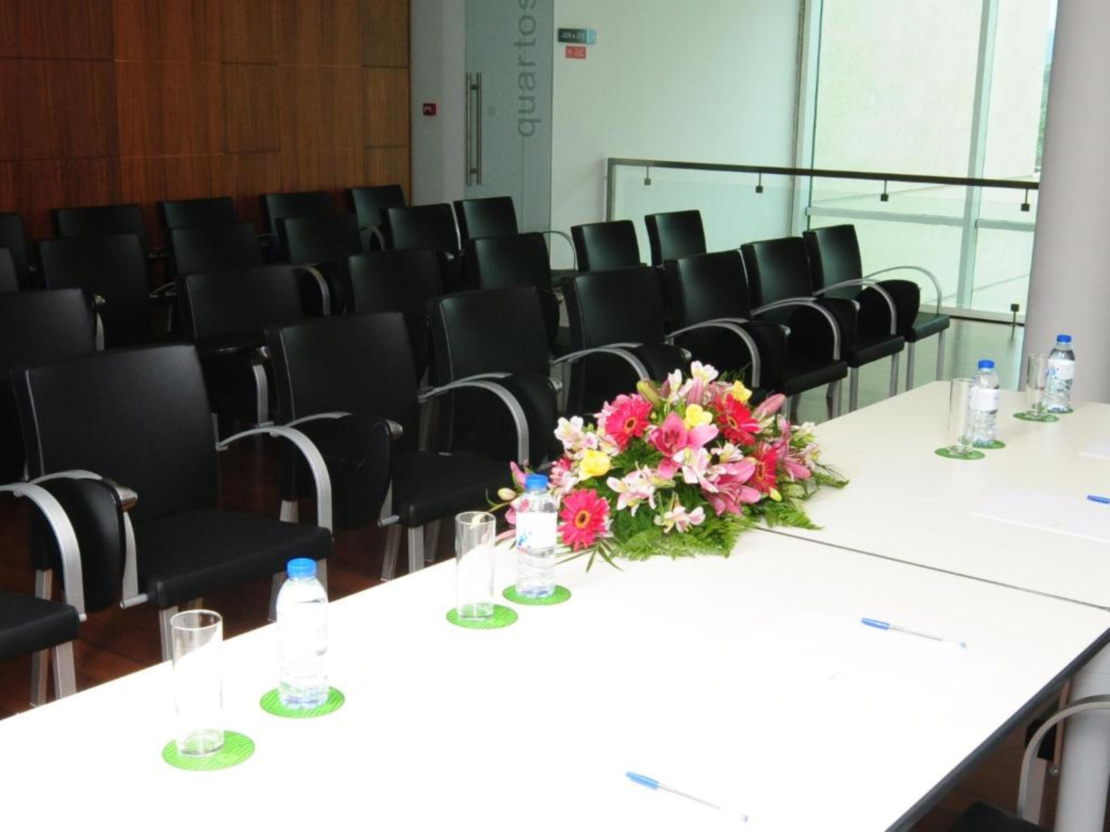 A conference room with rows of black chairs, a white table with a floral centerpiece, water bottles, and pens.