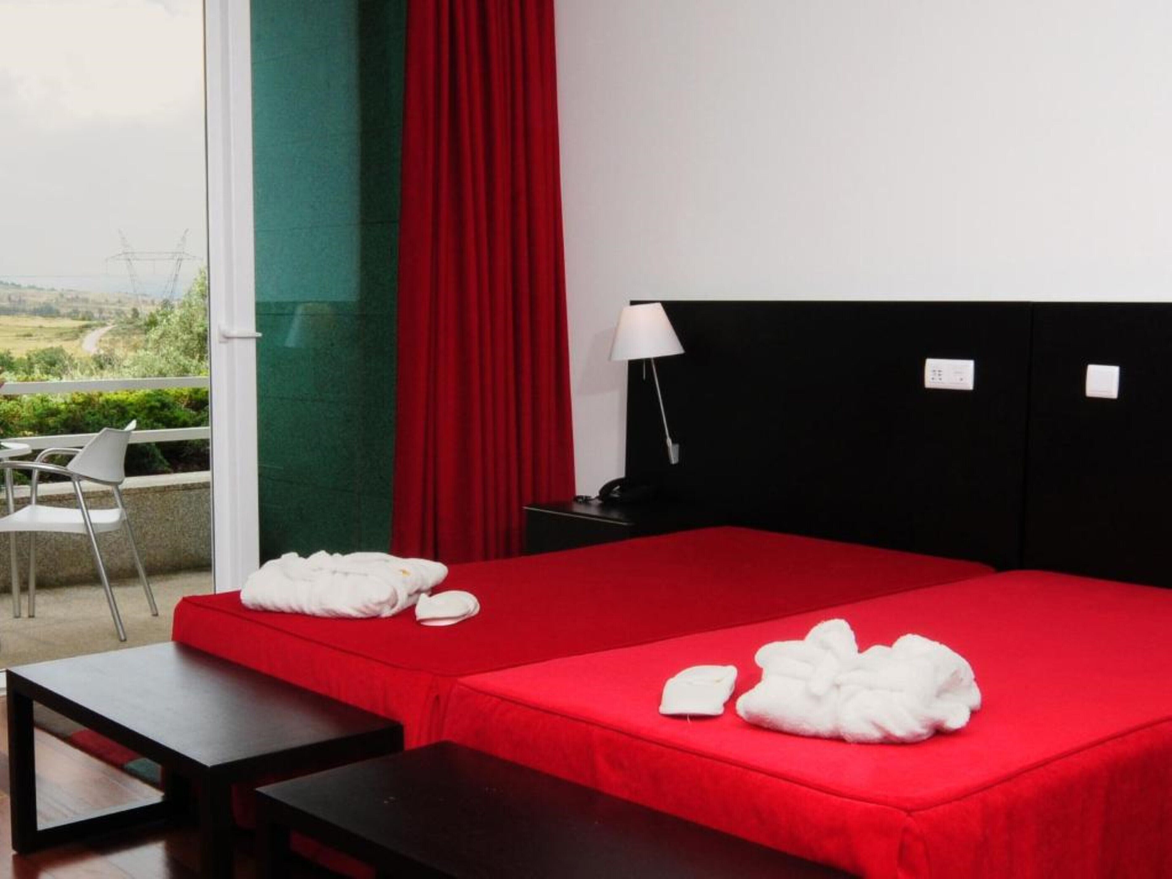Une chambre d’hôtel avec une couvre-lit rouge, deux serviettes blanches, une table de chevet noire avec une lampe, et une vue sur la verdure par la fenêtre.