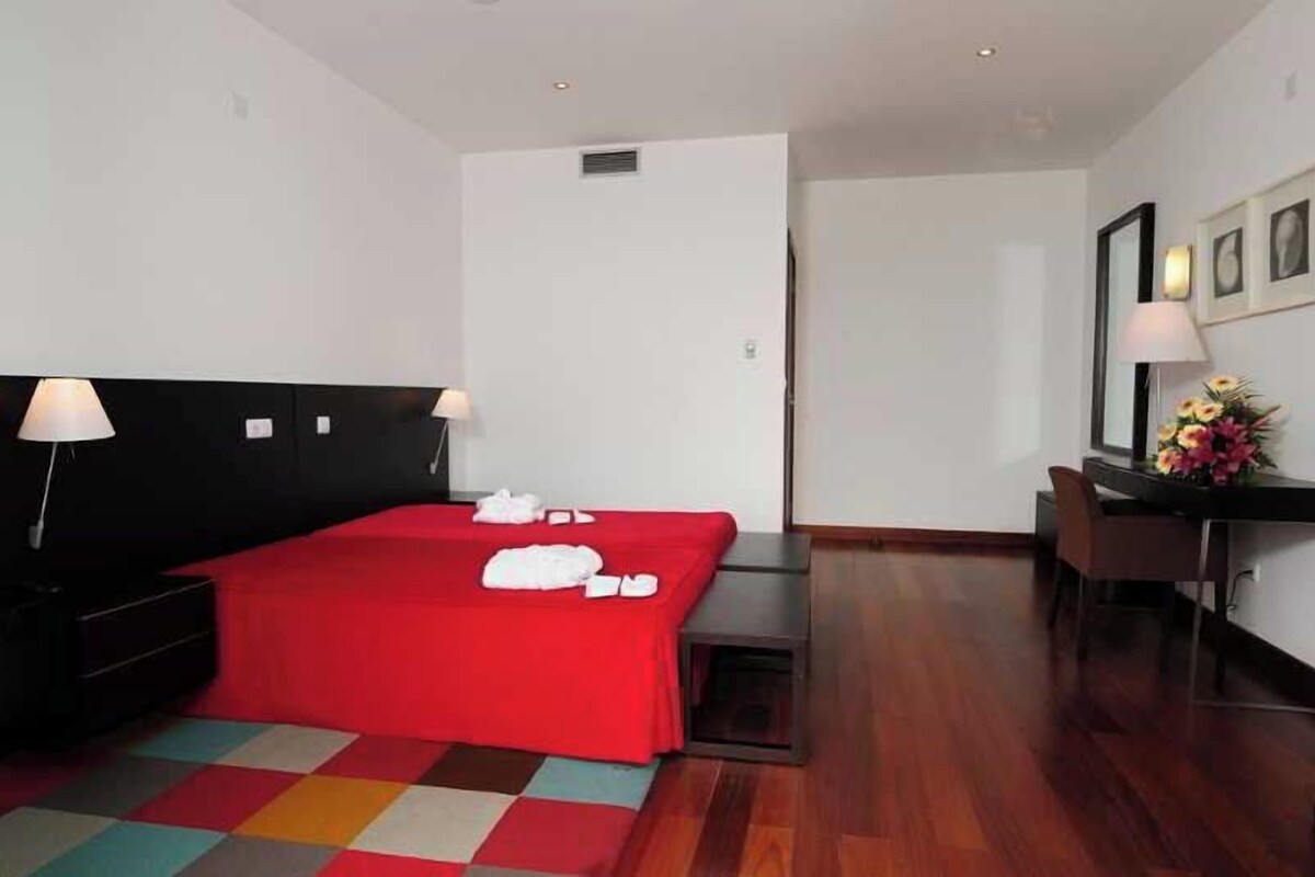 A hotel room with a red bedspread, a desk with a chair, a small table, and a vase of flowers.