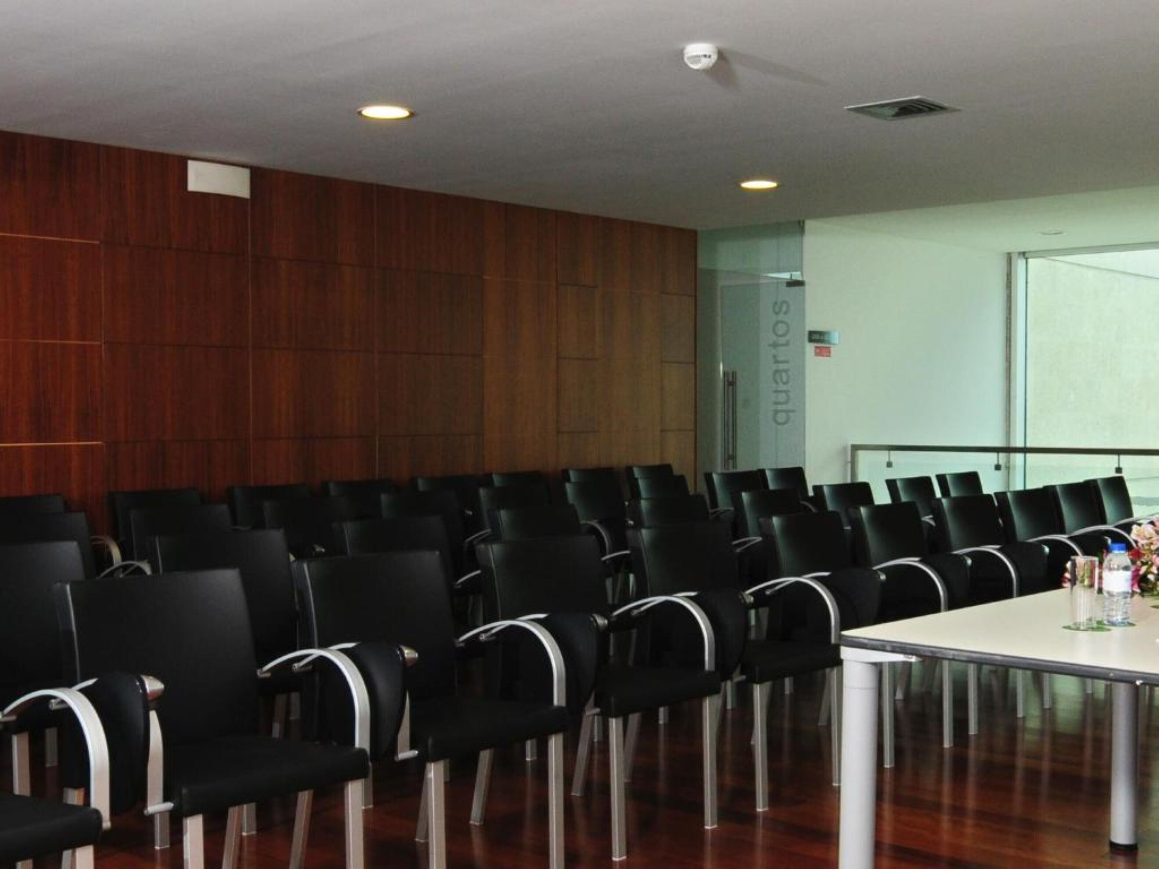 Une salle de conférence avec des rangées de chaises noires, une table blanche et des panneaux de bois sur les murs.
