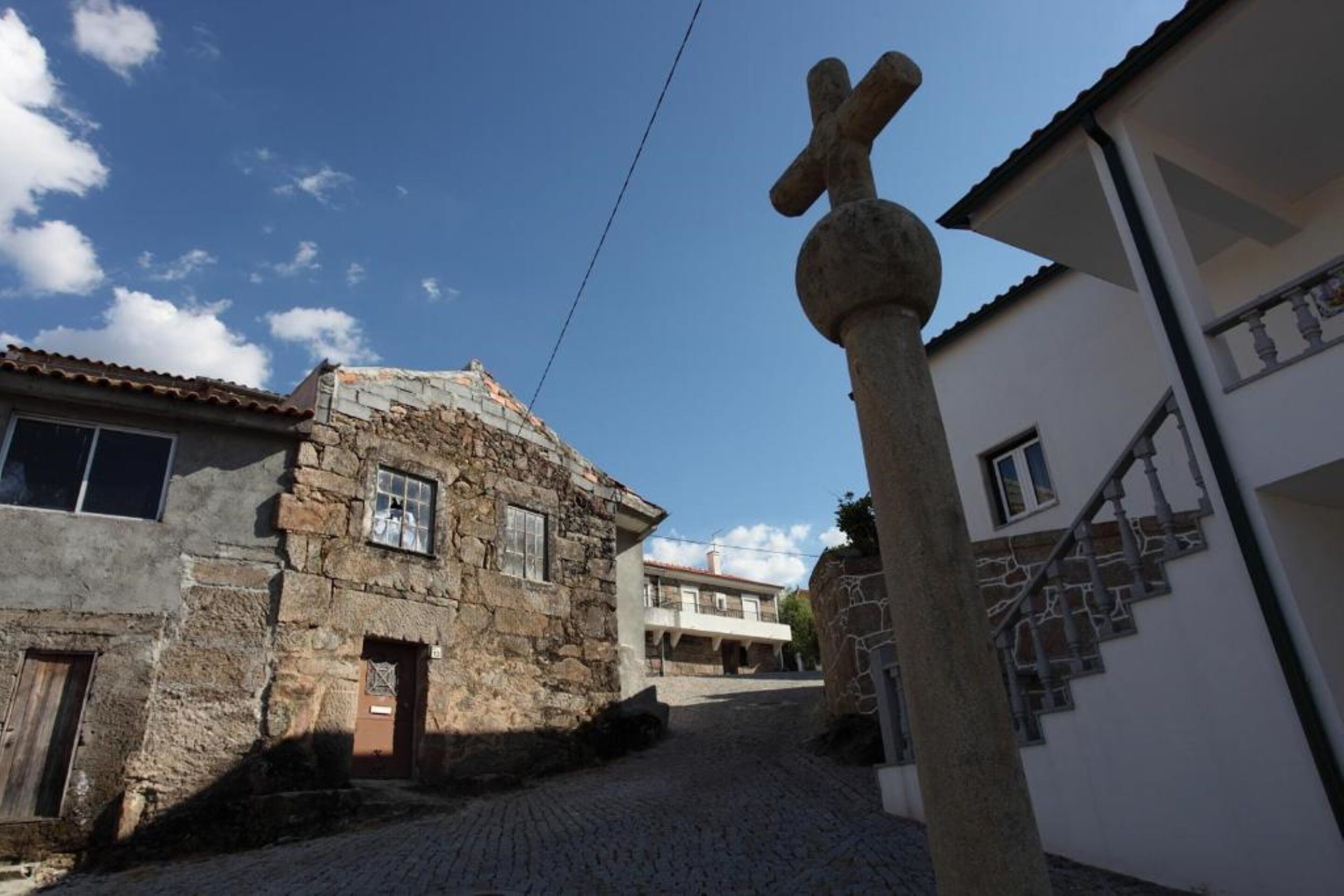 Une rue pavée bordée de bâtiments en pierre, avec une croix de pierre et un escalier menant à un bâtiment blanc doté d’un balcon.