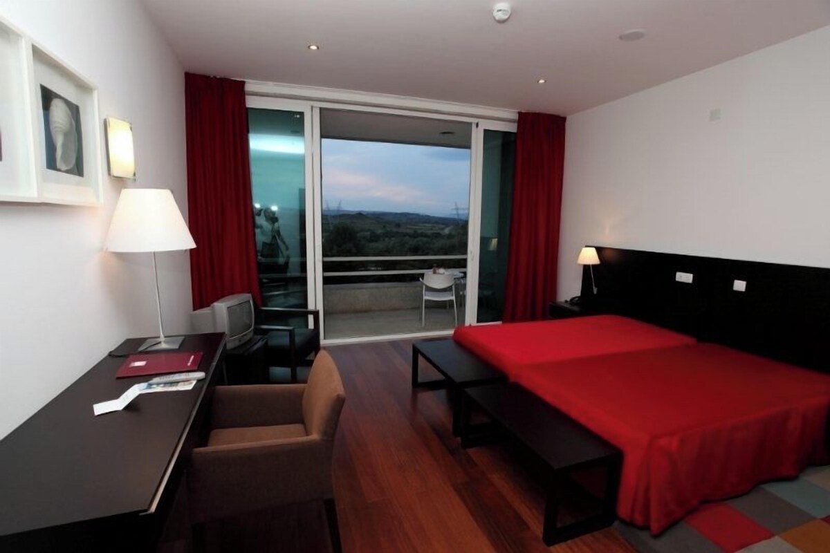 A hotel room with a bed, desk, chair, and a view through a sliding glass door.