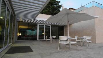Un espace extérieur aménagé en terrasse, avec des chaises blanches et un grand parasol, une entrée en verre et un mur en pierre.