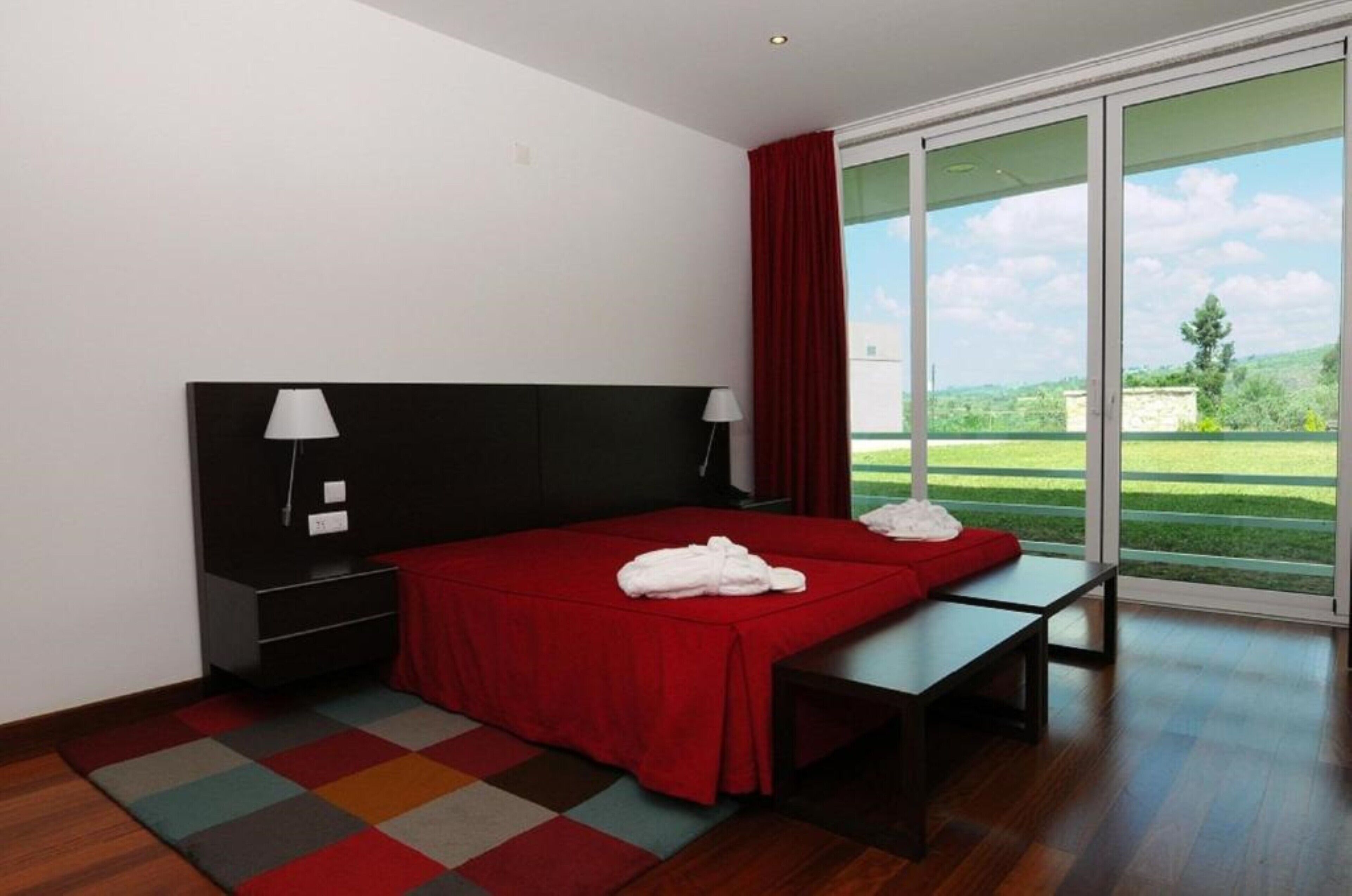 Una habitación de hotel moderna con una cama grande, una mesita de noche, una lámpara y vistas a un paisaje verde a través de una puerta corrediza de vidrio.
