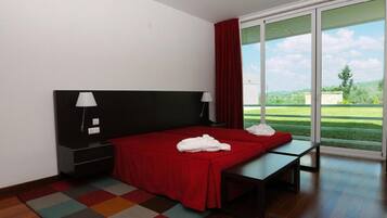 Una habitación de hotel moderna con una cama grande, una mesita de noche, una lámpara y vistas a un paisaje verde a través de una puerta corrediza de vidrio.