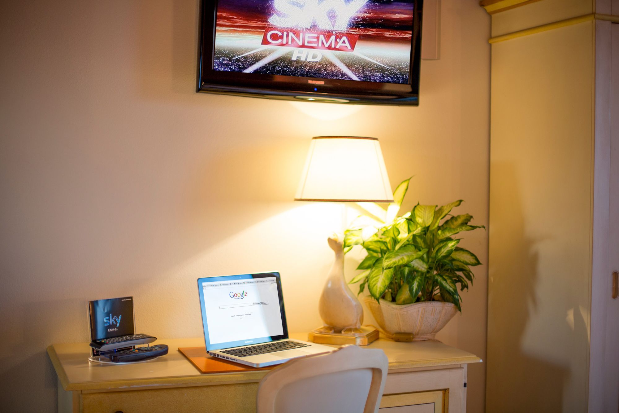 Habitación de hotel con una televisión mostrando una película, una laptop sobre un escritorio, una lámpara y una planta en maceta.