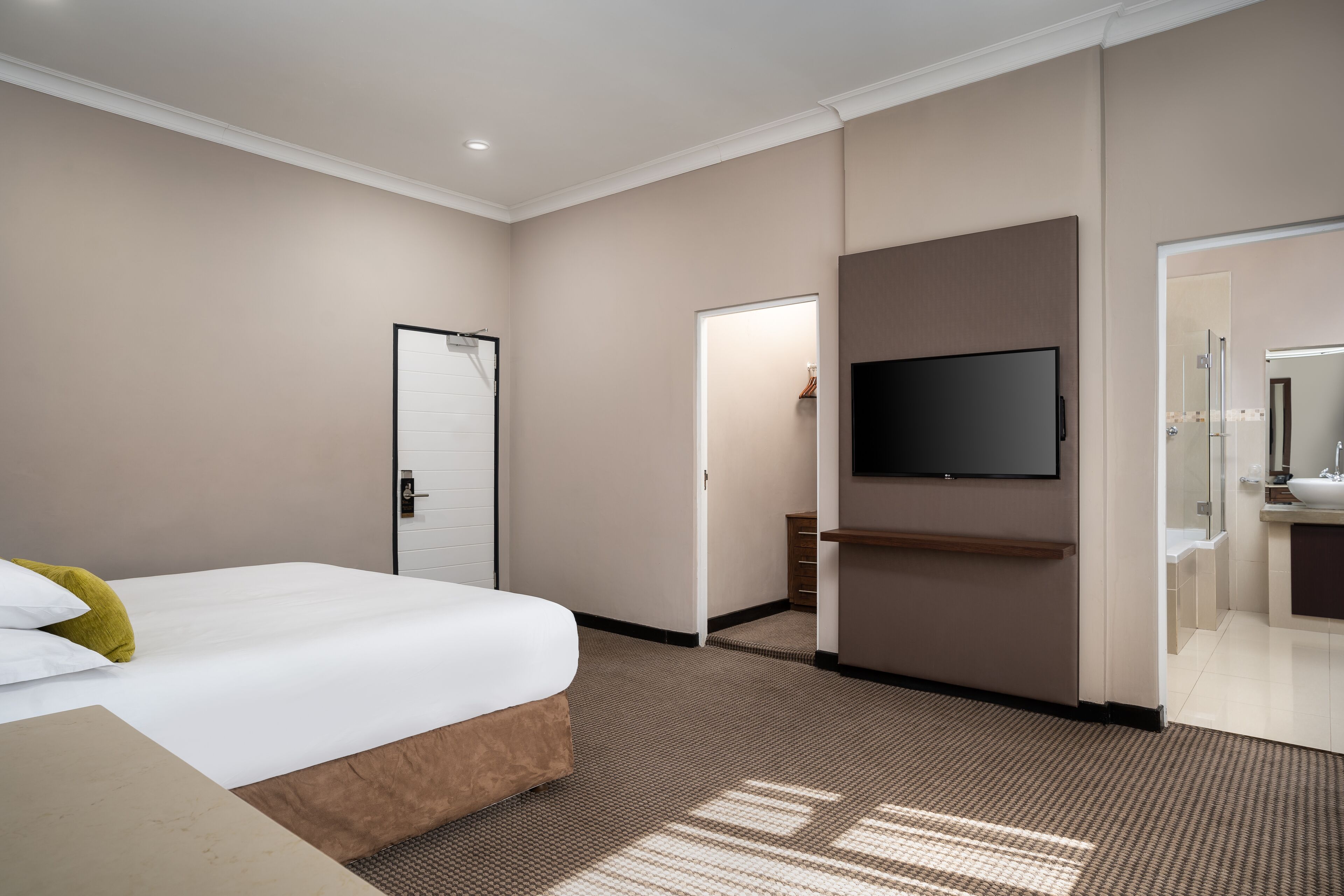 A hotel room with a bed, a TV mounted on the wall, a bathroom with a sink and mirror, and a door leading to another room.