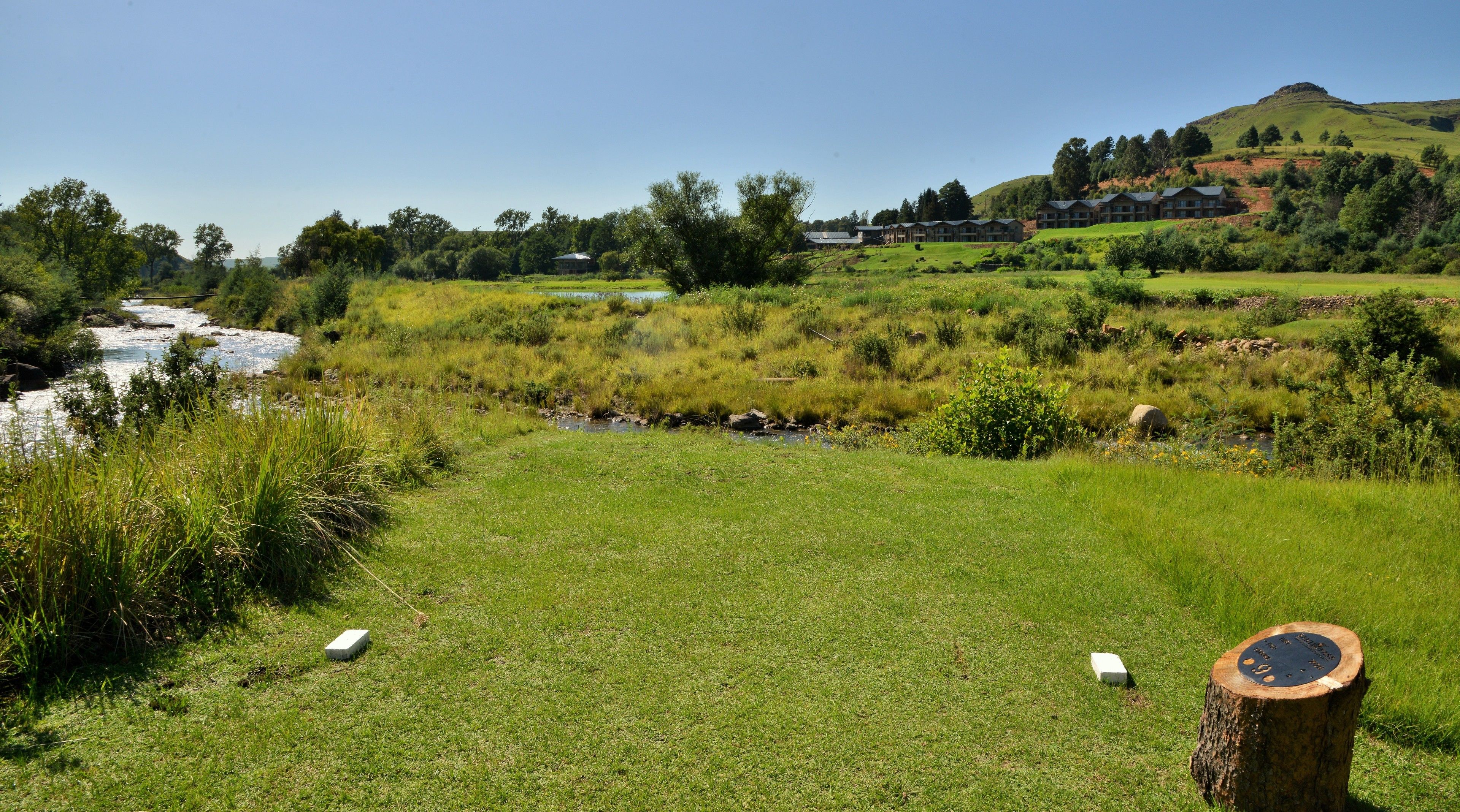 A grassy field with a small stream, a wooden post with a plaque, and a building complex in the background.