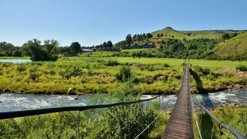 A wooden suspension bridge over a river, leading to a building with multiple rooms and a hill in the background.