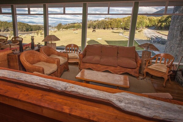 A cozy room with wicker furniture, a wooden bench, and a view of a grassy field with a mountain in the background.
