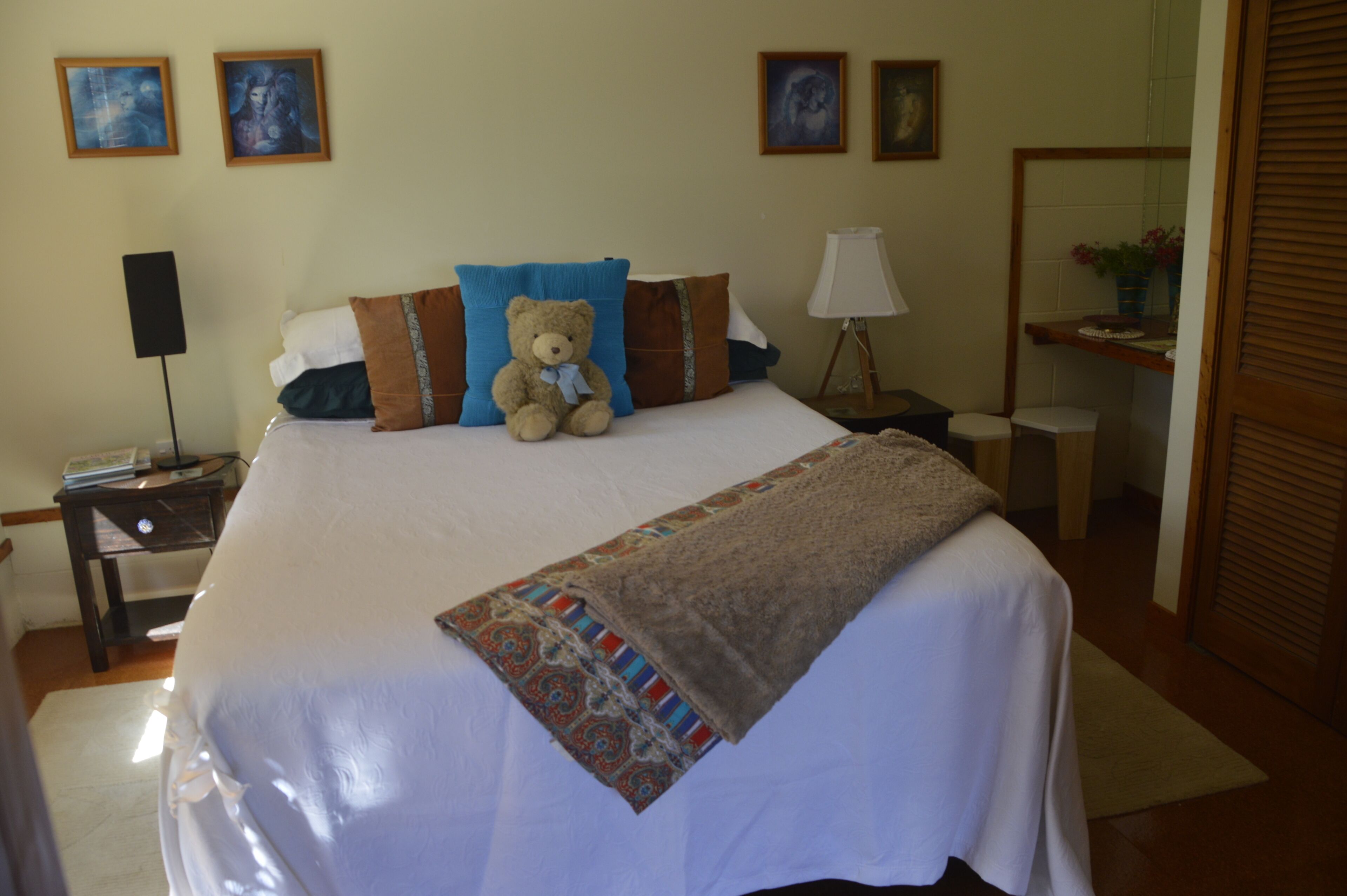 A neatly made bed with a teddy bear, pillows, and a blanket in a hotel room with artwork on the walls and a lamp on the nightstand.