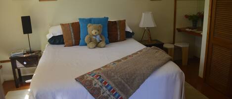 A neatly made bed with a teddy bear, pillows, and a blanket in a hotel room with artwork on the walls and a lamp on the nightstand.