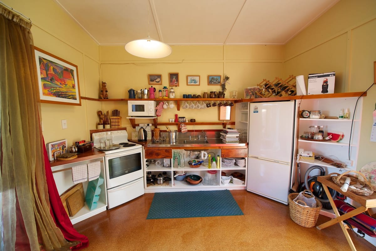 A compact kitchen with a microwave, stove, and refrigerator. There are shelves with dishes and a hanging rack for pots. A colorful painting is on the wall, and a blue mat is on the floor.