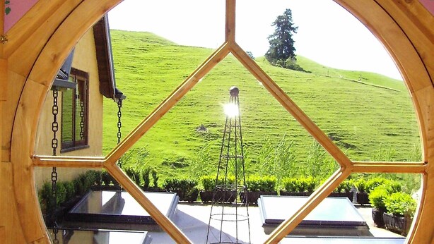 A circular window with a wooden frame and a view of a green hillside. - The Mudcastle (Upper Moutere)
