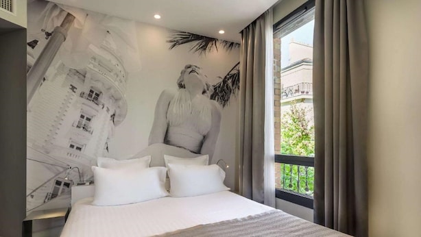 A hotel room with a large bed, a view of a building through the window, and a mural on the wall. - Hotel Sixteen Paris Montrouge (Montrouge)