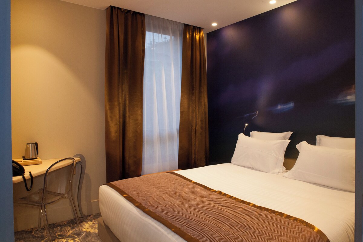 Quarto de hotel com cama, escrivaninha, cadeira e uma janela grande com cortinas.
